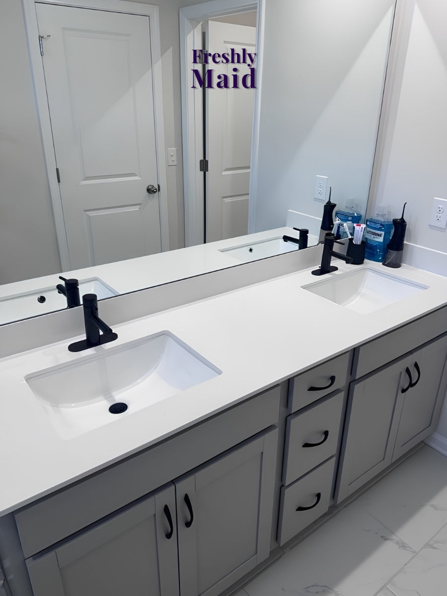 A double-sink bathroom vanity with a white countertop, white cabinets, and black faucets, shown in a real bathroom setting with a mirror, doors, and toiletries. A purple 'Freshly Maid' watermark appears near the top.