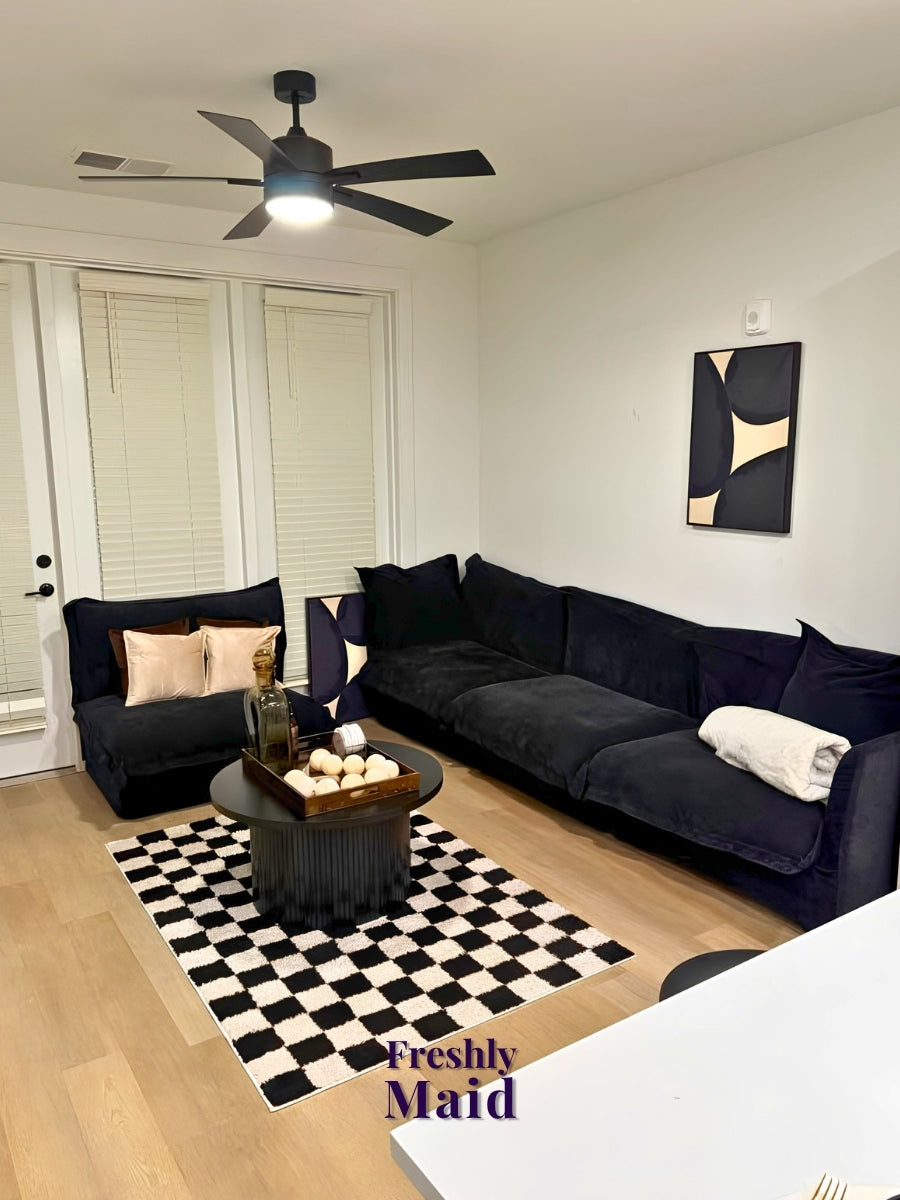 A modern living room setup featuring a dark navy blue velvet sectional sofa and matching armchair, styled with a black and white checkered rug, a round coffee table, and abstract wall art. A 'Freshly Maid' watermark is visible in the bottom right corner.
