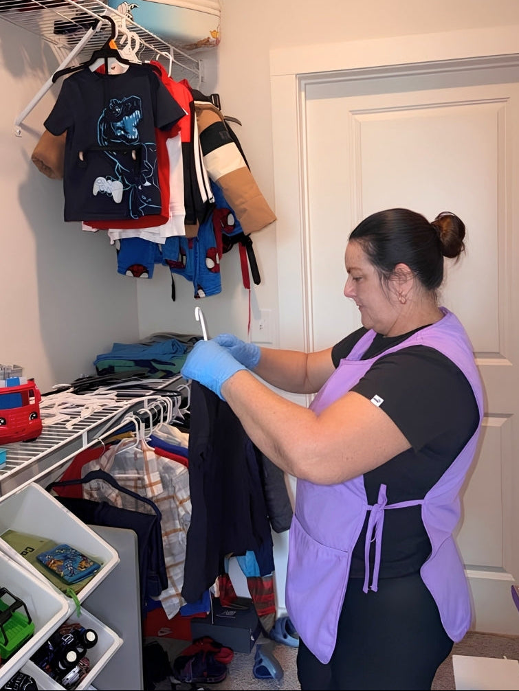 A woman wearing a purple and black apron over a black shirt stands in a closet organizing clothes on a white wire rack. The wall above her reads 'Freshly Maid'.
